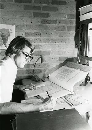 A young man in eyeglasses leans over a desk covered in papers and books. With his right hand he writes. The desk is against a brick wall. 